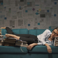 Stressed exhausted businesswoman sleeping on a couch at night, she is surrounded by piles of paperwork, overtime work and deadlines concept